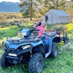 Two kids on an ATV near a family camping tent in a grassy field with mountains in the background.