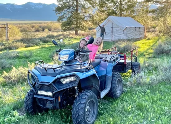 Two kids on an ATV near a family camping tent in a grassy field with mountains in the background.