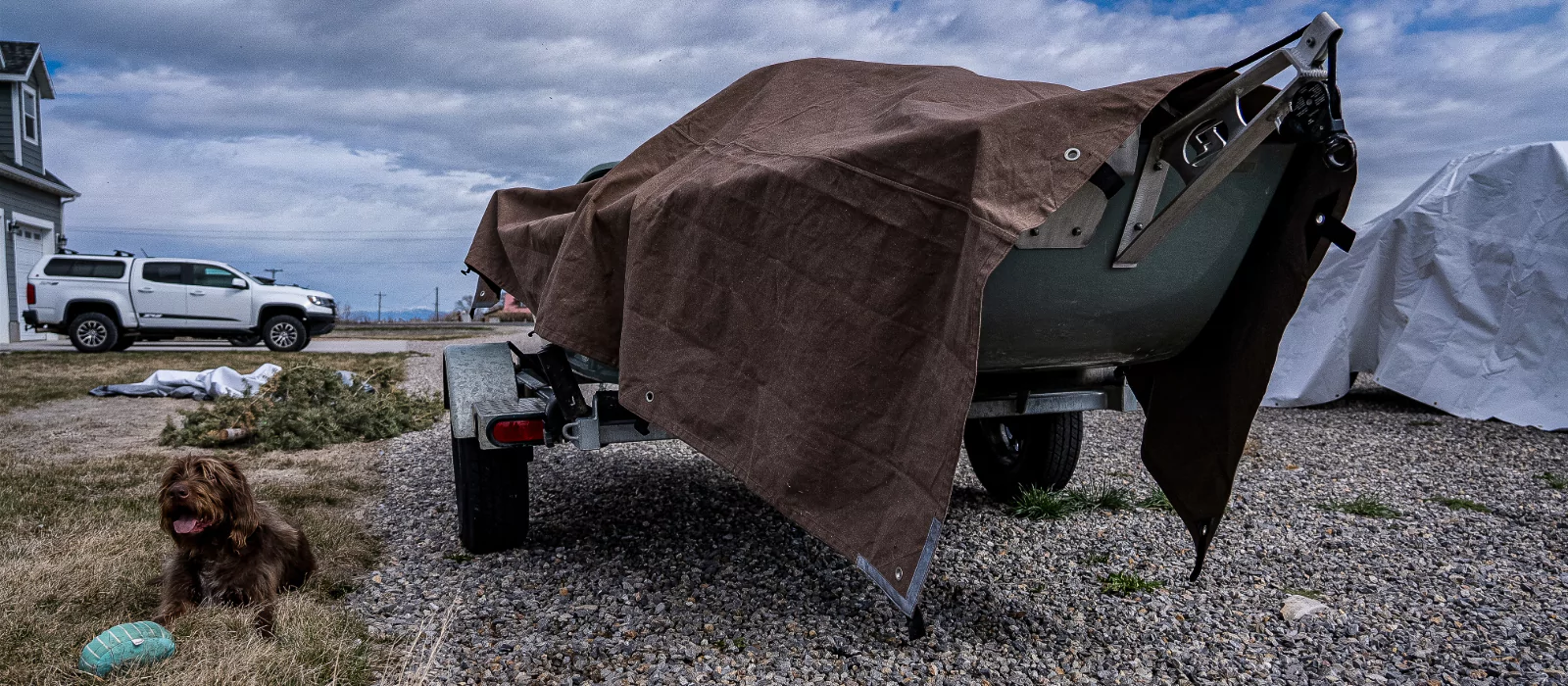 A dog rests next to a boat on a gravel road, with outdoor accessories concealed under traps nearby