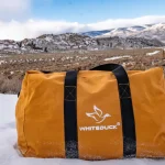orange canvas bag on snow covered ground with a mountainous landscape and cloudy sky in the background
