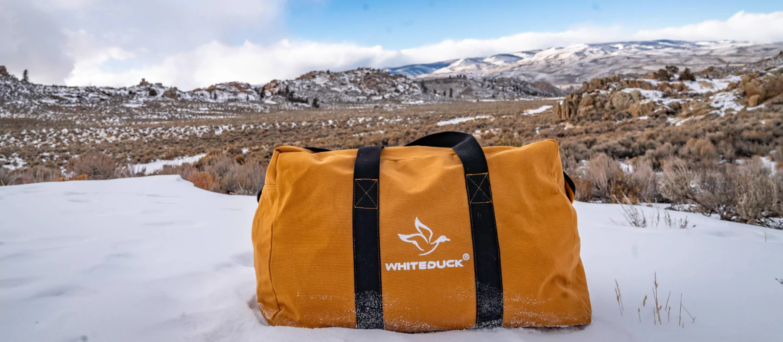 orange canvas bag on snow covered ground with a mountainous landscape and cloudy sky in the background