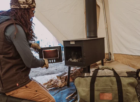 Camper setting up a wood-burning tent stove inside a canvas tent