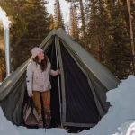 Woman enjoying winter camping in a snowy forest with a heated tent.