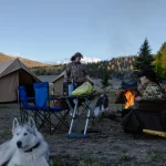 A family enjoying a camping trip in a forested area with tents, camping chairs, and a campfire, accompanied by a white dog, during early evening.