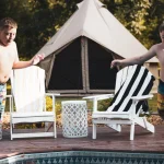 Two children in swimsuits jump into a pool from a wooden deck. A canvas tent and two chairs are visible in the background.