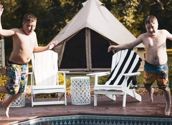 Two children in swimsuits jump into a pool from a wooden deck. A canvas tent and two chairs are visible in the background.