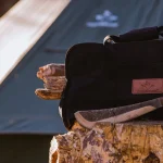 A durable log carrier bag from White Duck, filled with firewood, resting on a stump next to a camping tent and an axe