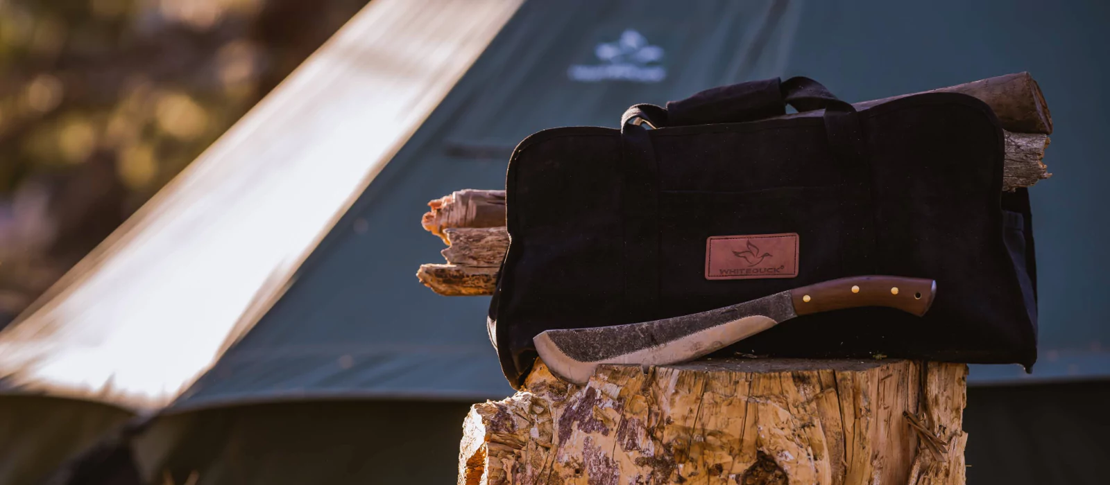 A durable log carrier bag from White Duck, filled with firewood, resting on a stump next to a camping tent and an axe