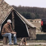 A man sits on a bench beside a tent alone, enjoying the outdoors in a serene natural setting