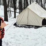 A man in a plaid shirt is positioned in the snow beside a canvas tent, with a serene winter scene in the background