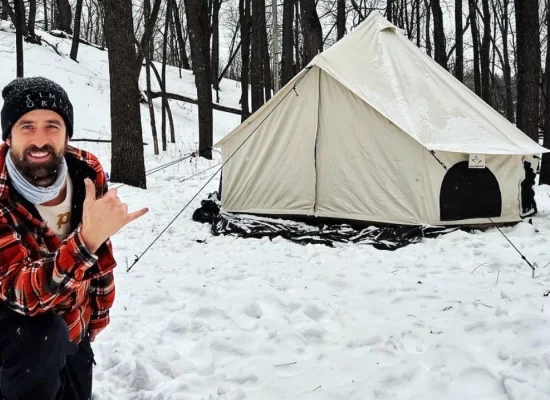 A man in a plaid shirt is positioned in the snow beside a canvas tent, with a serene winter scene in the background