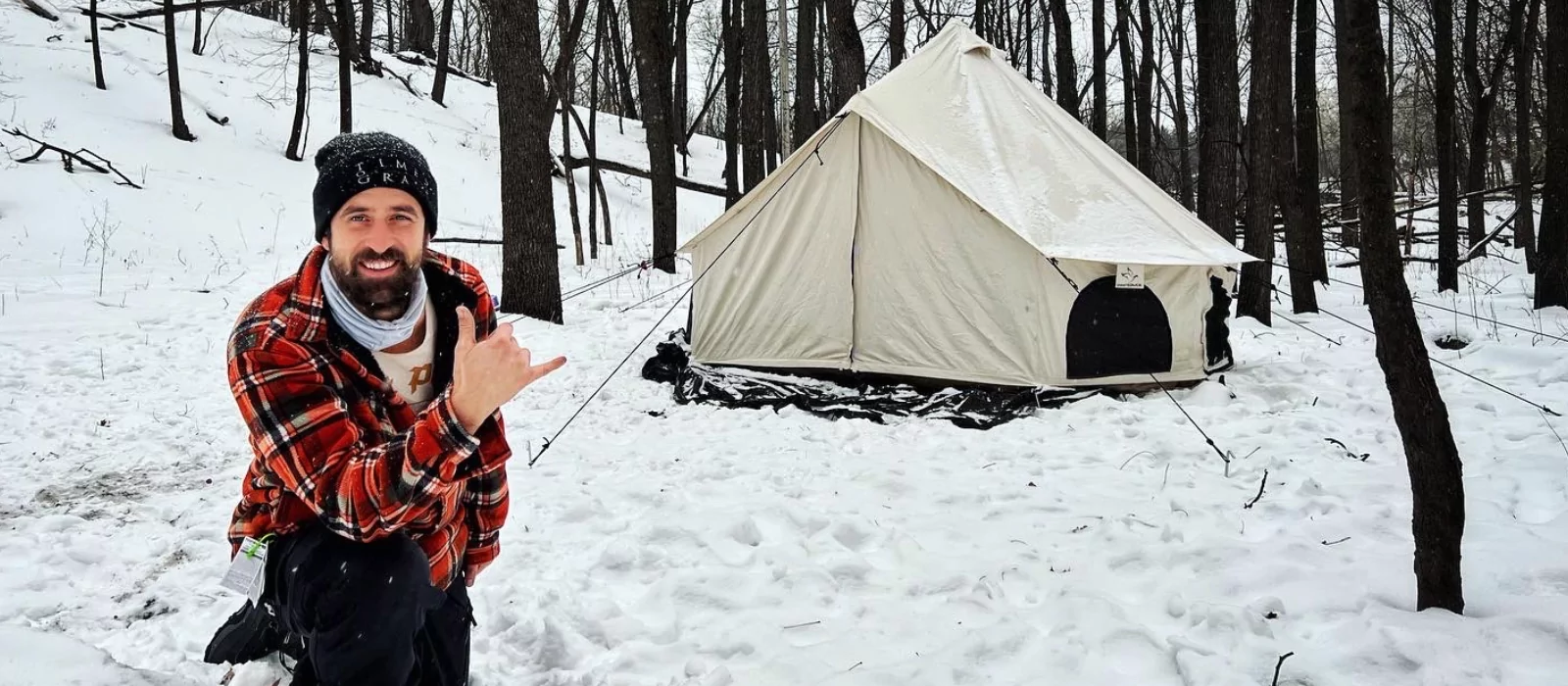 A man in a plaid shirt is positioned in the snow beside a canvas tent, with a serene winter scene in the background