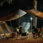 A nighttime camping scene with a tarp shelter, a lit lantern, and campers enjoying a warm meal by the fire.