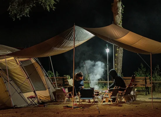 A nighttime camping scene with a tarp shelter, a lit lantern, and campers enjoying a warm meal by the fire.