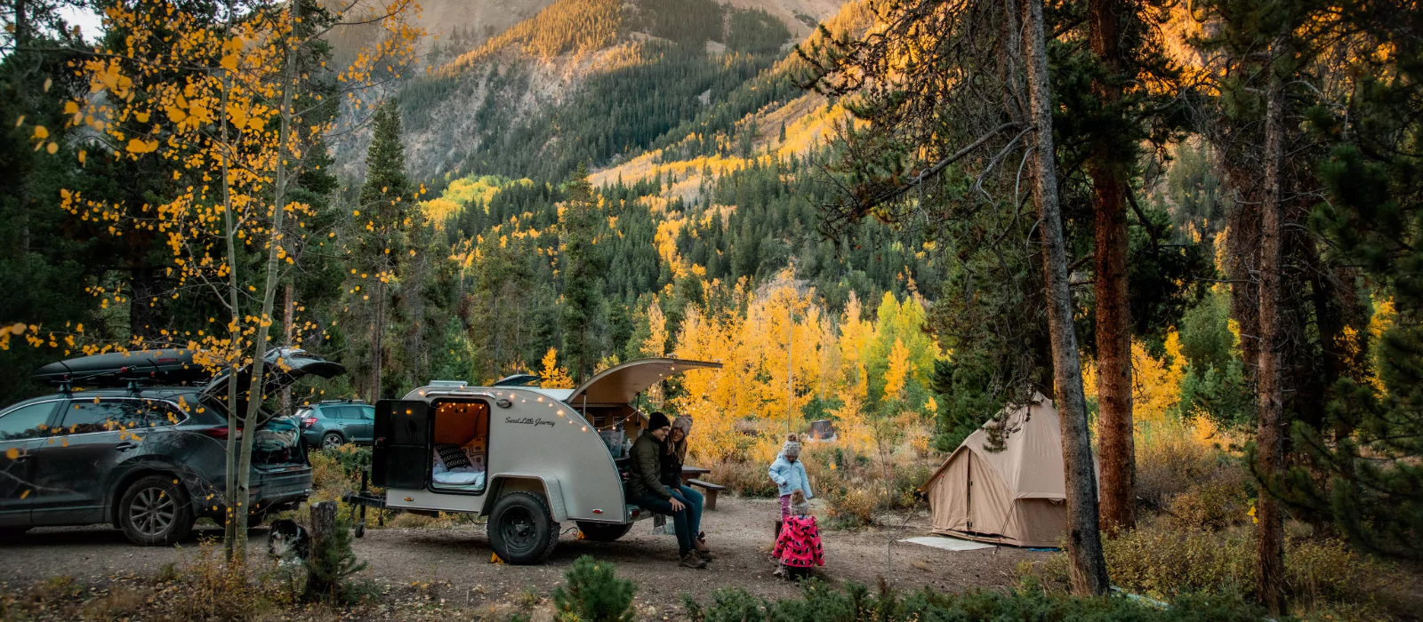 A cozy autumn camping scene in the mountains with a teardrop trailer, a canvas tent, and a family enjoying the outdoors among golden trees
