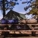 Best camping bags on a wooden picnic table with a tent and scenic mountain view in the background.