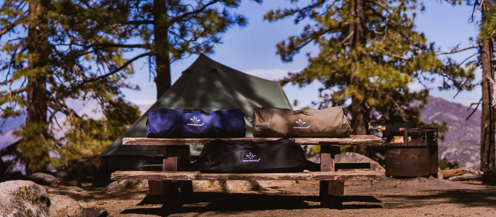 Best camping bags on a wooden picnic table with a tent and scenic mountain view in the background.
