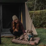 Camping bag beside a tent with a woman and her dog, showcasing essential outdoor gear for a perfect camping trip.
