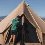 Man fixing the zipper of a camping tent in an open field with mountains and autumn trees in the background.