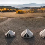A group of canvas tents set up in a spring field, with majestic mountains rising in the background