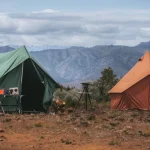 Two canvas camping tents set up in a remote mountain location with scenic views, outdoor gear, and a rugged wilderness backdrop.