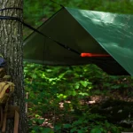 A bushcraft camping setup with a green tarp shelter tied between trees, a tactical backpack hanging on a tree, and camping gear in a forest.