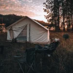A peaceful spring camping scene with a canvas tent set up in nature, surrounded by trees and mountains, as campers relax at sunset.