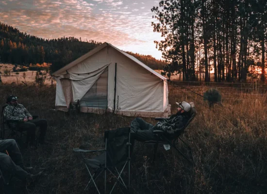 A peaceful spring camping scene with a canvas tent set up in nature, surrounded by trees and mountains, as campers relax at sunset.