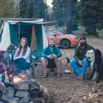 Family and dogs enjoying camping essentials like tents, fire pit, and outdoor gear at a forest campsite