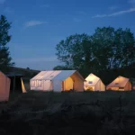 A scenic view of canvas tents illuminated from within at dusk, set in an outdoor campsite with trees and a darkening blue sky in the background.