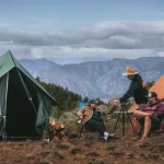 A family enjoying a spring camping trip in the mountains, sitting around a table near canvas tents with a scenic forest and mountain backdrop.