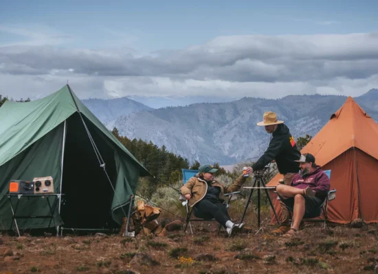 A family enjoying a spring camping trip in the mountains, sitting around a table near canvas tents with a scenic forest and mountain backdrop.