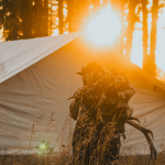 Hunter carrying gear at a summer hunting camp with a canvas wall tent during sunset in the forest