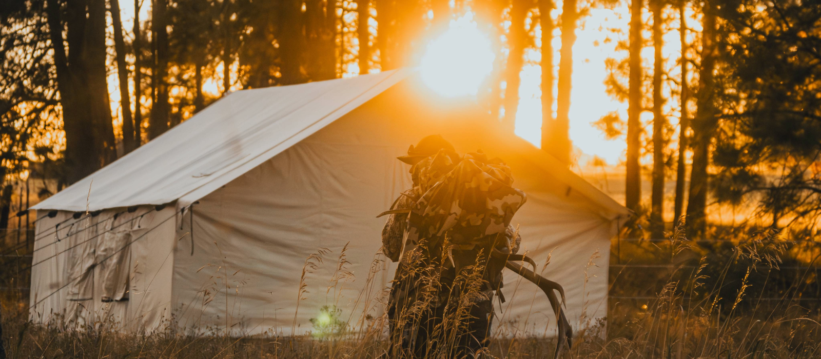 Hunter carrying gear at a summer hunting camp with a canvas wall tent during sunset in the forest