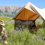 Woman enjoying spring camping surrounded by wildflowers with a canvas tent and mountain views in the background.