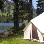 Canvas tent set up by a scenic lake during spring camping, surrounded by pine trees and mountains.