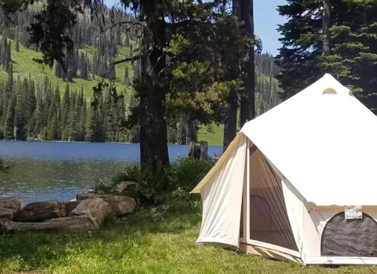 Canvas tent set up by a scenic lake during spring camping, surrounded by pine trees and mountains.