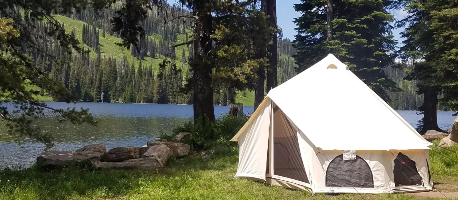 Canvas tent set up by a scenic lake during spring camping, surrounded by pine trees and mountains.