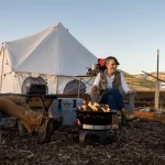 A family enjoying spring camping with kids around a campfire in front of a canvas tent in the mountains.