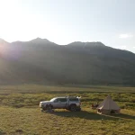 Scenic summer camping setup in the mountains with a canvas tent and SUV parked on an open grassy field at sunrise