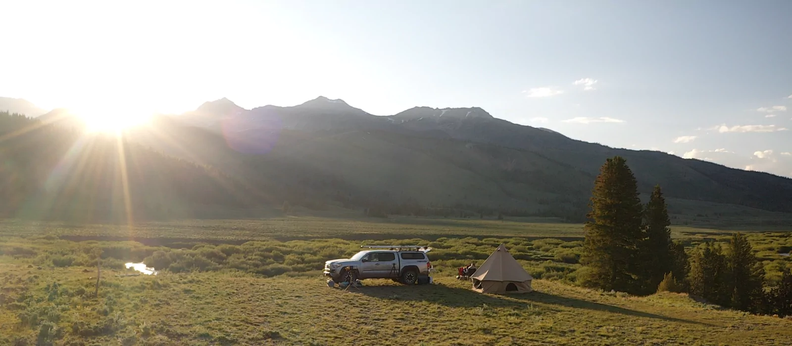 Scenic summer camping setup in the mountains with a canvas tent and SUV parked on an open grassy field at sunrise