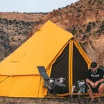 Couple setting up a bright yellow bell tent during summer camping in a rocky mountain landscape with a dog nearby.