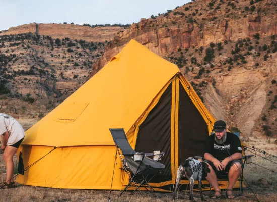 Couple setting up a bright yellow bell tent during summer camping in a rocky mountain landscape with a dog nearby.