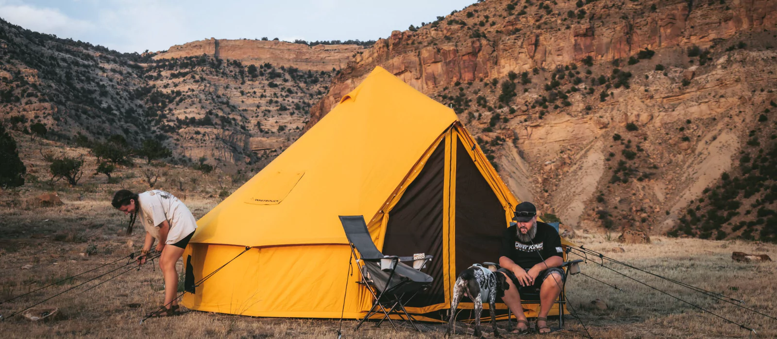 Couple setting up a bright yellow bell tent during summer camping in a rocky mountain landscape with a dog nearby.