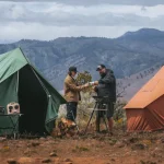 Two campers enjoying a budget camping trip with canvas tents set up in a scenic mountain landscape.