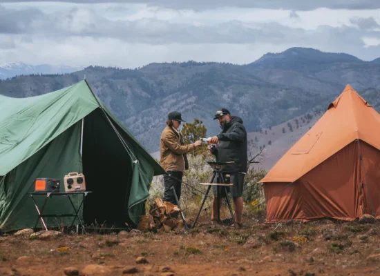 Two campers enjoying a budget camping trip with canvas tents set up in a scenic mountain landscape.