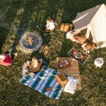 Campers enjoying a camp meal around a fire pit with tent and cooking setup on grassy field
