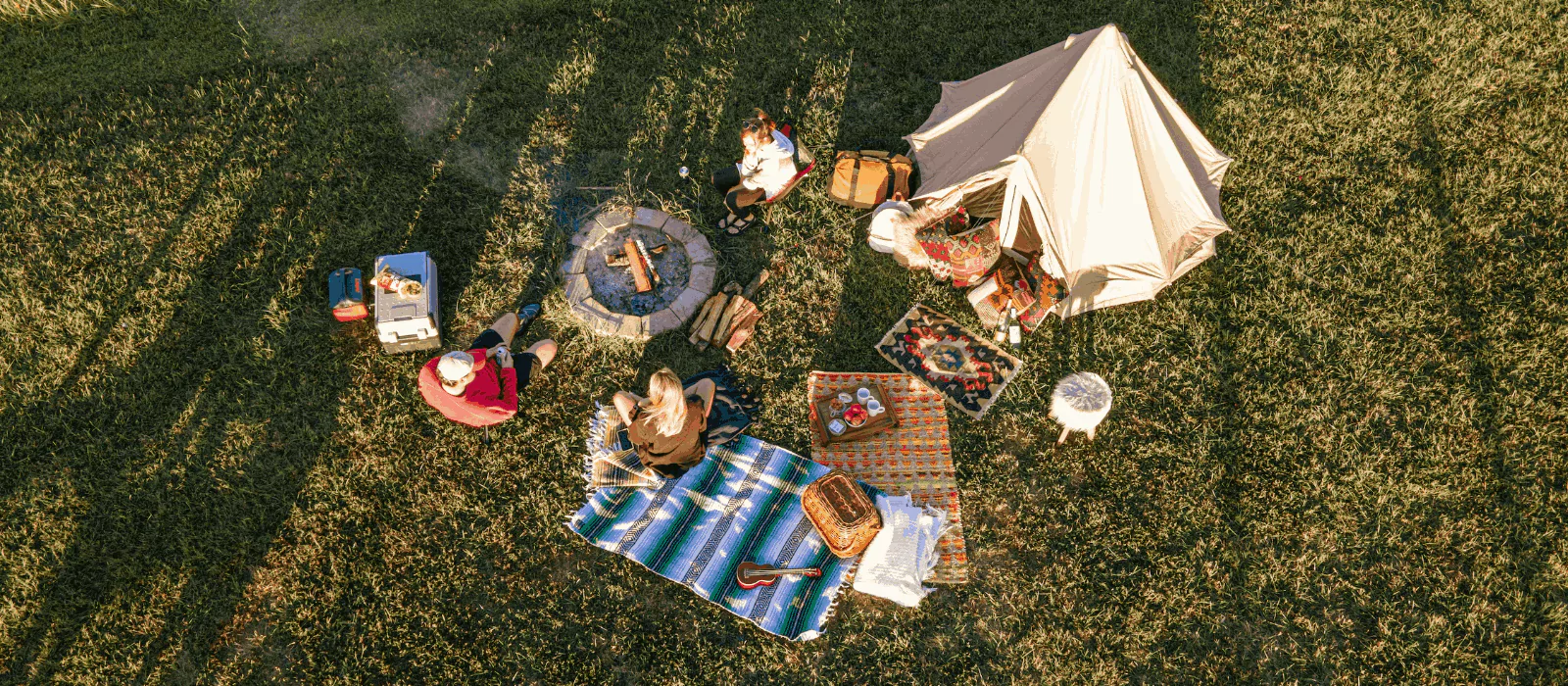 Campers enjoying a camp meal around a fire pit with tent and cooking setup on grassy field