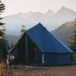 Woman and dog walking toward a tent at sunrise in a national park with mountain views