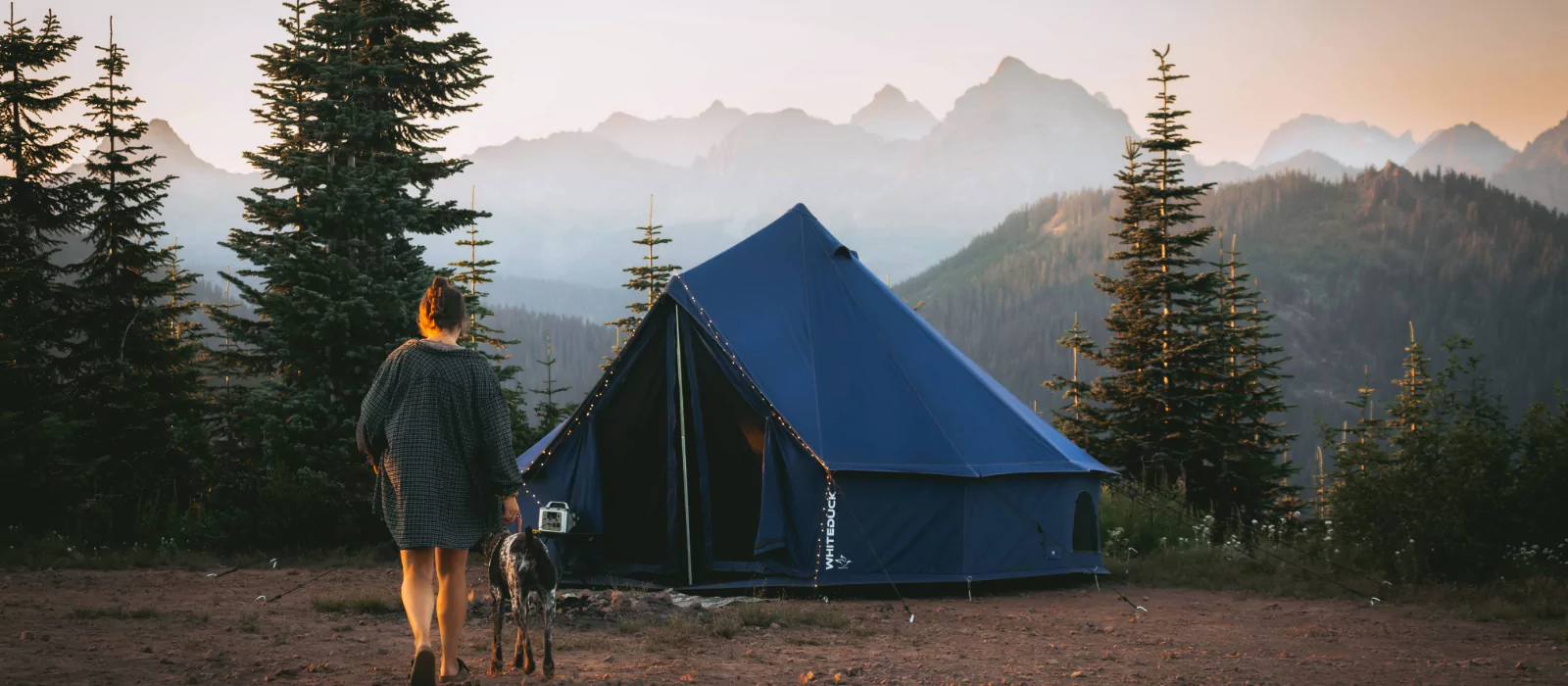 Woman and dog walking toward a tent at sunrise in a national park with mountain views